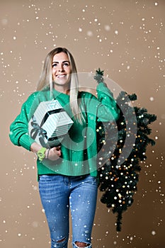 Beautiful attractive blond hair woman hold Christmas fir tree with bokeh lights and gift box in green sweater
