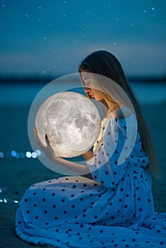 Beautiful attractive girl on a night beach with sand and stars hugs the moon, Artistic Photography