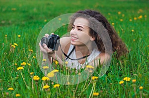 Beautiful, attractive girl-photographer with curly hair holds a camera and lying on the grass with blooming dandelions