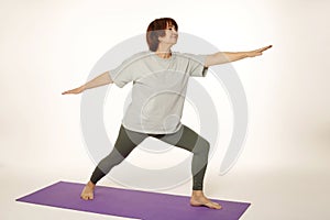 Beautiful attractive photogenic woman of 60s practicing yoga on mat in studio warrior pose