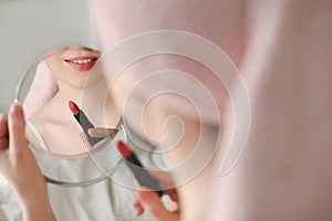 Beautiful attractive smiling woman paints lips with red lipstick using a small round mirror during home makeup in the