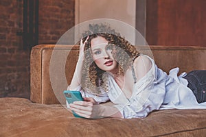 Beautiful attractive woman with curly hair lies on brown sofa and searching the net using her mobile phone