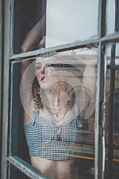 Beautiful attractive woman with curly hait and grey top staying in the window and looking