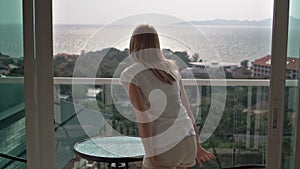 Beautiful attractive woman opens sliding-doors to balcony. Goes outside and sit on a chair relaxing.