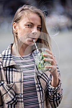 Beautiful attractive young caucasian woman or twentysomething girl drinking cold mojito with straw in city center during photo