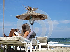 Beautiful Attractive Young Girl with Long Hair Sitting Relaxing and Reading Book under Straw Beach Parasol Umbrella and Blue Sky