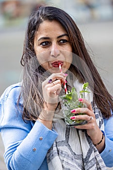 Beautiful attractive young indian woman or twentysomething girl drinking cold mojito with straw in city center during photo