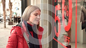 Beautiful attractive young woman looking at shop window. Red winter coat. Sales signs. Evening night.