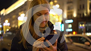 Beautiful attractive young woman standing in a street and using her smartphone. City lights Cars.