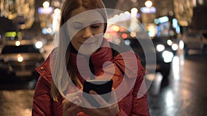 Beautiful attractive young woman standing in a street and using her smartphone. City lights Cars.