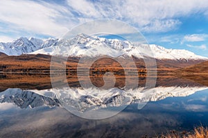 Beautiful autumn mountain lake and mountains. Reflection of snowy peaks and red coniferous taiga on the surface of a mountain lake