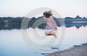 The beautiful baby with curly hair cheerfully jumps on the bank of the lake. Active spring walk in the open air