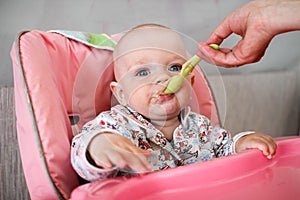 Beautiful baby eats porridge from mom's hand. He is sitting on a pink children' chair.