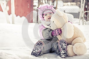 Beautiful baby embracing toy plush bear sitting on snow in park in cold sunny winter day during winter holidays