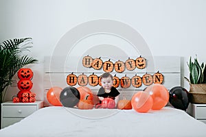 Beautiful baby girl in halloween costume at home, sitting on bed with Halloween decoration, Lifestyle indoors