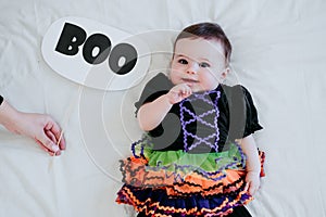 Beautiful baby girl with halloween costume at home, sitting on bed with Halloween decoration, mother hand holding BOO sign.