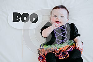 Beautiful baby girl with halloween costume at home, sitting on bed with Halloween decoration, mother hand holding BOO sign.