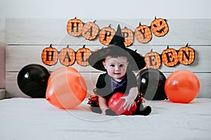 Beautiful baby girl in witch halloween costume at home, sitting on bed with Halloween decoration, Lifestyle indoors