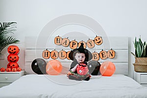 Beautiful baby girl in witch halloween costume at home, sitting on bed with Halloween decoration, Lifestyle indoors