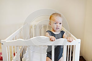 Beautiful baby standing in crib. Portrait of cute baby girl stand in cot and looking around. Toddler learning to stand in bed at