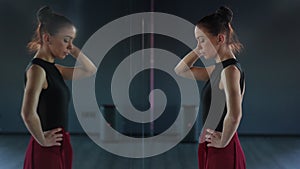 A beautiful ballerina stands and thinks while looking at her reflection in the mirror of the choreographic hall.