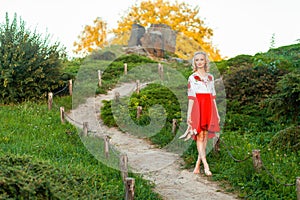 Beautiful barefoot woman in stylish red white dress holding shoes in hand and walking on path at hill. standing on road village