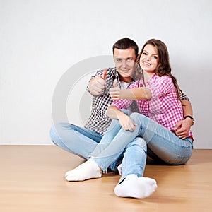 Beautiful barefoot young couple in casual jeans sitting leaning against a white wall on the wooden floor in the living room