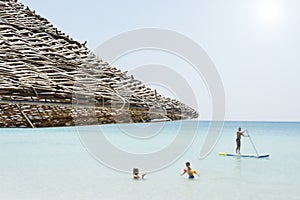 Beautiful beach resort with straw umbrellas on a blue sky and white clouds. On the background a man is surfing standing up with a