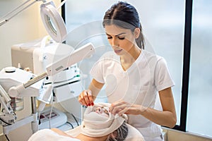 Beautiful beautician doing beauty treatment, covering female client& x27;s face with towel in beauty salon.