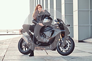 A beautiful biker girl leaning on her superbike outside a building.