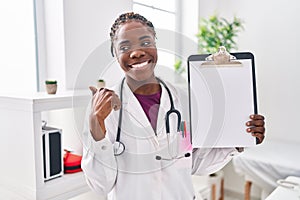 Beautiful black doctor woman holding clipboard pointing thumb up to the side smiling happy with open mouth