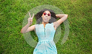Beautiful black girl lying on green grass, showing peace gesture and sticking out her tongue. Top view