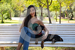 Beautiful black haired woman is sitting with her doberman puppy on a white bench in the park. The woman enjoys the company of her