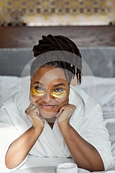 beautiful black lady in bathrobe after shower puts gold patches under eyes and use cosmetics