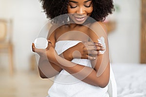Beautiful black lady wearing towel after bath, applying cream, rubbing in nourishing body butter at home
