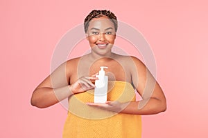 Beautiful black obese woman wrapped in towel holding bottle with moisturising body lotion, lady using skincare product