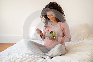 Beautiful black pregnant woman sitting on bed and holding bowl of salad