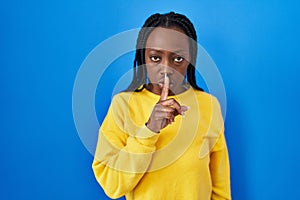 Beautiful black woman standing over blue background asking to be quiet with finger on lips
