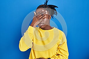 Beautiful black woman standing over blue background covering eyes with hand, looking serious and sad