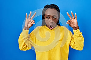 Beautiful black woman standing over blue background looking surprised and shocked doing ok approval symbol with fingers