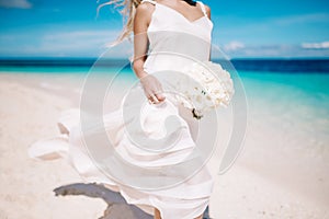 Beautiful blond bride in white wedding dress with white wedding bouquet stand on sea shore. Tropical turquois sea on the backgroun