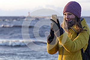 Beautiful blond caucasian girl in winter hat and bright coat looking at her mobile phone at the sunset seaside