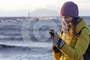 Beautiful blond caucasian girl in winter hat and bright coat looking at her mobile phone at the sunset seaside