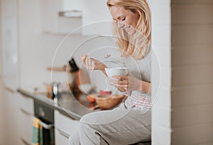 Beautiful blond caucasian woman posing in her kitchen, while drinking coffee or tea and eating a healthy breakfast meal full of ce