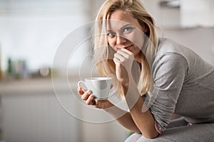 Beautiful blond caucasian woman posing in her kitchen, while drinking coffee or tea and eating a healthy breakfast meal full of ce