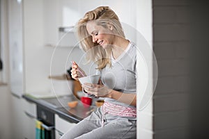 Beautiful blond caucasian woman posing in her kitchen, while drinking coffee or tea and eating a healthy breakfast meal full of ce