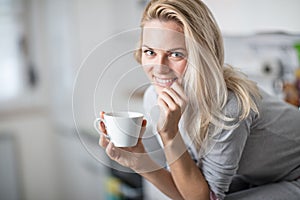 Beautiful blond caucasian woman posing in her kitchen, while drinking coffee or tea and eating a healthy breakfast meal full of ce