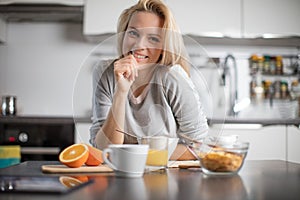 Beautiful blond caucasian woman posing in her kitchen, while drinking coffee or tea and eating a healthy breakfast meal full of ce