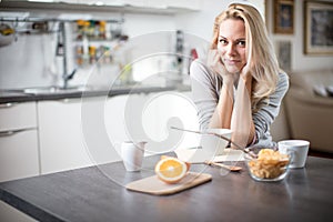 Beautiful blond caucasian woman posing in her kitchen, while drinking coffee or tea and eating a healthy breakfast meal full of ce