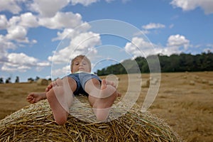 Beautiful blond child, boy, lying on a haystack in the field. Amazing landscape, rural scene with clouds, tree and empty road